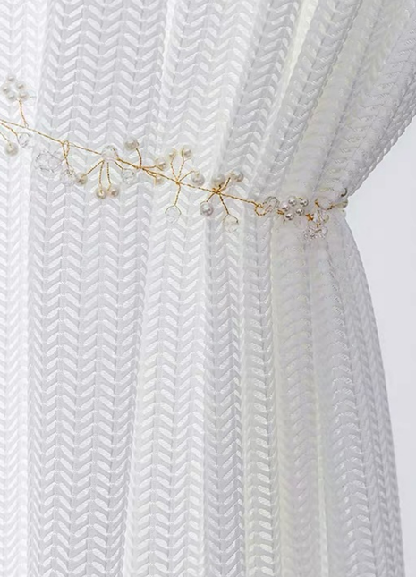 A close-up view of a white, sheer curtain with a decorative gold-colored ribbon or chain attached to it.