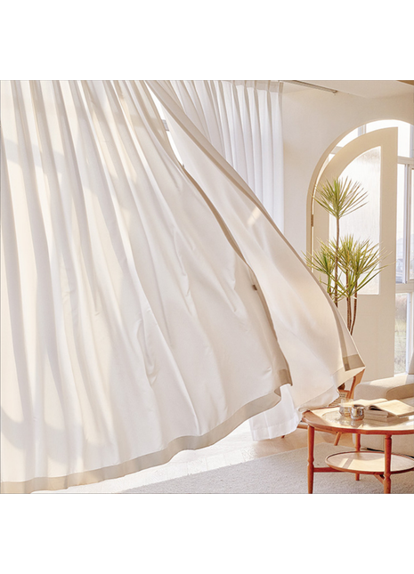 a room with a large window, white curtains blowing in the wind, and a wooden table with a glass vase and a book on it.