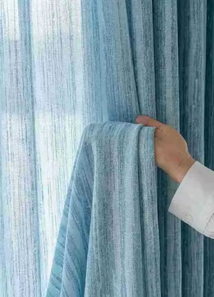 Rideau bleu ciel au tissage strié écarté à la main devant un voilage assorti