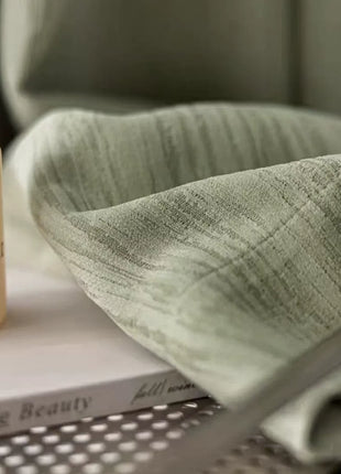 Pan de rideau vert chenille replié sur un livre et un flacon, ambiance douce et naturelle